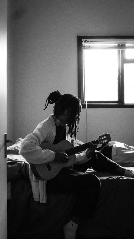 Black-and-white frame — the man seated with his guitar, expression over distraction